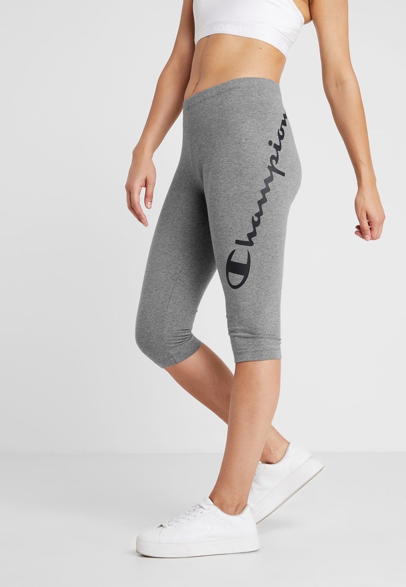 Szare, krótkie legginsy wykonane z mieszanki bawełny, z czarnym logo "Champion" nadrukowanym pionowo wzdłuż lewej nogawki, w połączeniu z białymi sneakersami.
