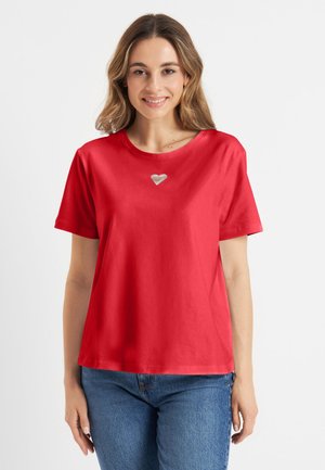 Kobieta z włosami do ramion uśmiechająca się, ubrana w czerwoną koszulkę z małym białym sercem na piersi oraz niebieskie dżinsy.