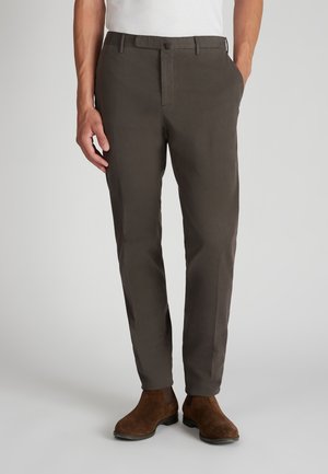 Man in een donkergrijze slim-fit broek en bruine suède Chelsea boots, staand met een hand in de zak tegen een effen achtergrond.