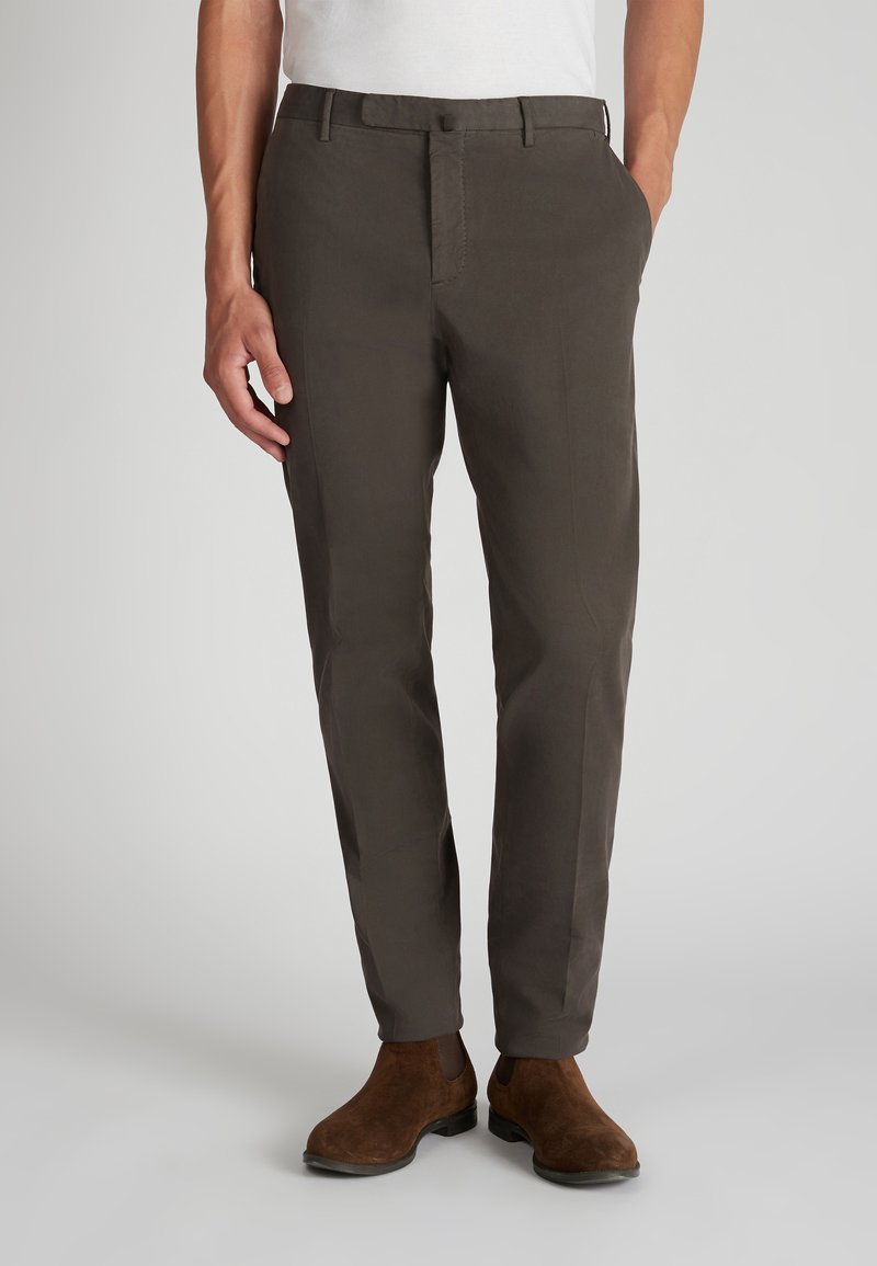 Hombre con pantalones oscuros de corte slim y botas Chelsea de ante marrón, de pie con una mano en el bolsillo contra un fondo liso.