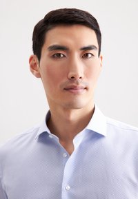 Giovane uomo con capelli corti e scuri, che indossa una camicia azzurro chiaro a motivi, che guarda dritto di fronte a uno sfondo leggero e semplice.
