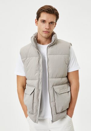 Chaleco acolchado gris con cuello alto, cierre frontal y dos bolsillos en el pecho. Llevado sobre una camiseta blanca, presenta una textura suave y ligera.