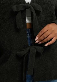 Svart texturerad kofta med två knutna rosetter fram, bärs över en vit topp och kombineras med blå denim. Tyget verkar mjukt och mysigt.