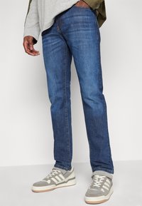 Dark blue denim jeans with a straight fit, featuring subtle fading and stitching. Paired with grey and white sneakers.