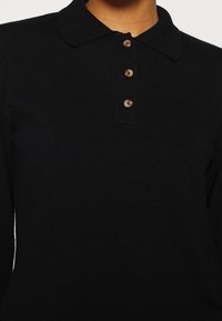 Polo à manches longues noir en maille avec trois boutons marron et un col doux porté par une personne visible du cou jusqu'au milieu du torse.