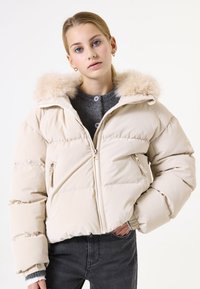Beige Daunenjacke mit einem von Fell gefütterten Kapuze, gestepptem Design, Reißverschlusstaschen und elastischen Bündchen. Getragen über einem grauen Pullover.