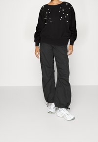 Felpa nera con borchie decorative, vestibilità rilassata e polsini a coste, abbinata a pantaloni cargo larghi e scuri e sneakers bianche.