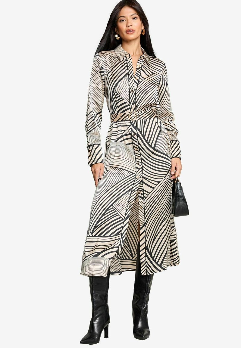 Abito beige a maniche lunghe con bottoni, con strisce geometriche nere, cintato in vita, presenta uno spacco ed è abbinato a stivali neri fino al ginocchio.