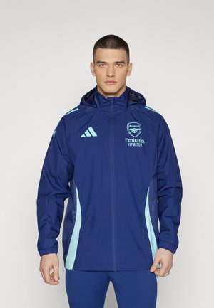 Chaqueta impermeable de color azul marino con acentos en azul claro, cierre de cremallera y capucha. Presenta el logo de Arsenal y la marca "Emirates" en la parte frontal.