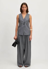 Gilet gris sans manches avec un décolleté en V profond et trois boutons noirs, associé à un pantalon gris à jambes larges. Modèle tenant une pochette noire.
