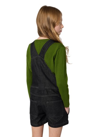 Zwarte denim dungarees met verstelbare banden, gecombineerd met een groene longsleeve trui. Opvallend detail op de achterzak, zachte textuur en contrasterende stiksels.