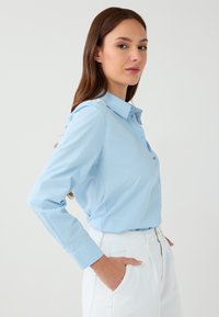 Light blue button-up shirt with a structured collar, long sleeves, and a smooth texture. Paired with white trousers featuring side pockets.
