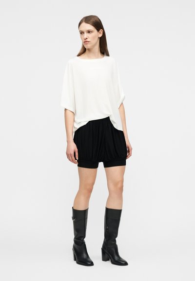 Γυναίκα που στέκεται φορώντας λευκό άνετο κοντομάνικο τοπ, μαύρες σορτς με ελαστική μέση και μαύρες μπότες με τακούνι μέχρι το μέσο της γάμπας, σε απλό φόντο.