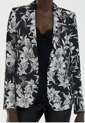 Schwarz-weiß gemusterter Blazer mit floralem Design, getragen über einem schwarzen Top mit Spitzenbesatz, kombiniert mit schwarzen Hosen.