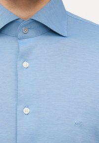 Ανοιχτό μπλε υφασμάτινο πουκάμισο με κουμπιά και μυτερό γιακά, με μικρό κεντημένο λογότυπο "MK" στο στήθος.