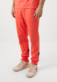 Herre joggingbukser i koralfarve i glat stof med en slim fit, med sorte striber ved anklerne. Sammen med lyse sneakers.