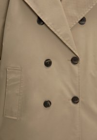 Veste beige croisée en tissu lisse, dotée de larges boutons noirs et d'une poche latérale unique.