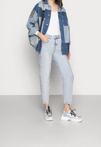 Kvinna i en patchwork denimjacka, vit topp, ljusa jeans, vita och blå sneakers, och bär en grå axelväska.