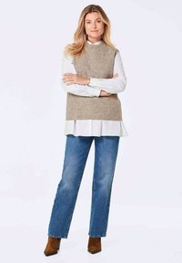 Un pull en tricot beige sans manches porté sur une chemise blanche à col, associé à un jean droit bleu et des bottes à chevilles marron. Design simple et décontracté.