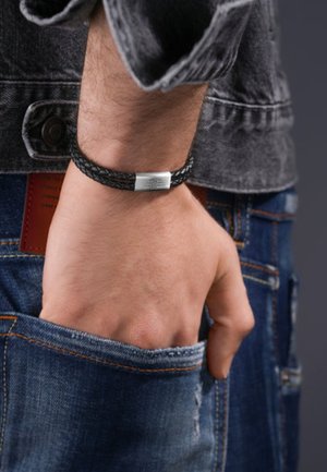 Bracelet en cuir tressé noir avec un fermoir en métal argenté, porté au poignet, complété par un jean en denim avec des accents décolorés.