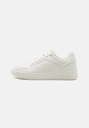 Witte lage Champion sneakers met veters, het kenmerkende "C" logo aan de zijkant, en een getextureerde rubberen zool op een eenvoudige achtergrond.
