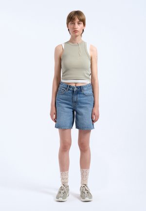 Hellgrünes ärmelloses Top und blaue Denim-Shorts. Das Model trägt beigefarbene Spitzenstrümpfe und graue Sneakers. Einfaches Design ohne sichtbare Muster.