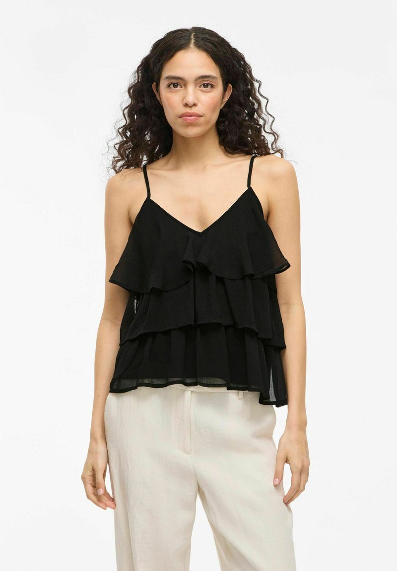 Camisola nera a strati con sottili spalline regolabili, avente strati arricciati e scollatura a V, realizzata in tessuto leggero. Sfondo semplice.