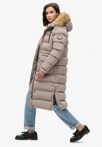 Long manteau rembourré de couleur beige clair avec une capuche doublée de fourrure, présentée avec une fermeture éclair, des fentes latérales et un badge sur la manche gauche. Porté au-dessus de jeans clairs et de chaussures noires.