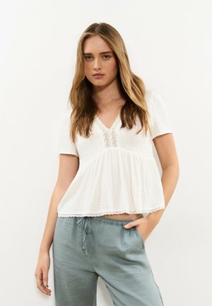 Mujer joven con cabello largo y rubio, vestida con una blusa blanca de manga corta con detalles de encaje y pantalones sueltos azul claro con cordón ajustable.