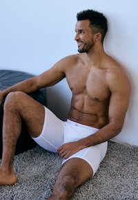Caleçons boxers blancs avec une bande d'accent rouge, fabriqués en matière extensible. Modèle assis contre un mur sur un tapis gris.