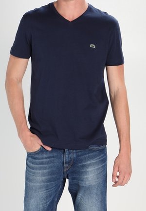Homme portant un t-shirt bleu marine à col en V avec un petit logo crocodile vert et un jean bleu, une main dans la poche, debout devant un fond uni.