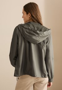 Giacca leggera verde oliva con cappuccio, dalla texture liscia, polsini elasticizzati e orlo cintato. Presenta una schiena ventilata per la traspirabilità.