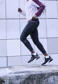Collants de sport noirs avec des accents violets, portés avec une veste blanche et bordeaux et des baskets noires, en train de courir sur une surface en pierre.