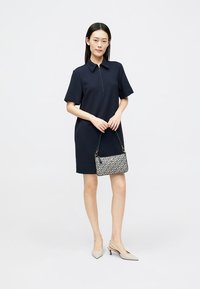 Vestido polo azul-marinho com gola com zip, mangas curtas, combinado com uma bolsa estampada em branco e azul-marinho. O modelo usa sapatos de salto nude com bico fino.