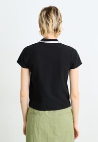 Čierna polo košeľa s krátkym rukávom a bielym golierom, so ribbed textúrou a zakriveným lemom. Nosená s zelenou sukňou.