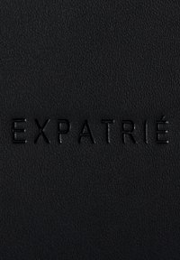 Černý texturovaný materiál s embosovaným nápisem "EXPATRIÉ." Povrch má jemný zrnitý vzor a matný povrch.