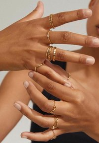 Gold rings in various styles, including twisted, smooth, and accented with a small stone, worn on multiple fingers. Skin tone is warm.