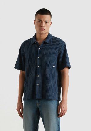 Mann trägt ein marineblaues, strukturiertes Kurzarm-Hemd mit Knopfleiste und Brusttasche, kombiniert mit hellblauen Jeans, steht vor weißem Hintergrund.