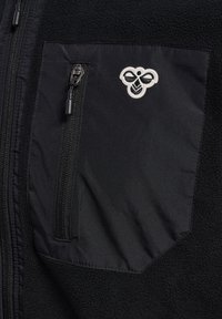 Black fleece jacket featuring a zippered pocket with an embroidered bee logo, contrasting fabric textures, and a smooth zipper pull.