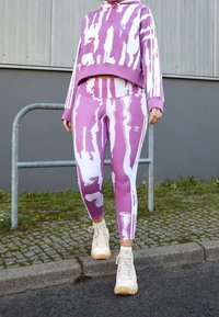 Fialová a bílá vzorovaná mikina s cropped designem; odpovídající fialové legíny se bočními pruhy a abstraktními bílými akcenty. Krémové tenisky.