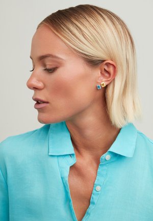Quadratische Ohrstecker mit blauen und gelben Steinen, die in goldfarbenem Metall gefasst sind. Tragen von einem Model in einem hellblauen Hemd mit Kragen.