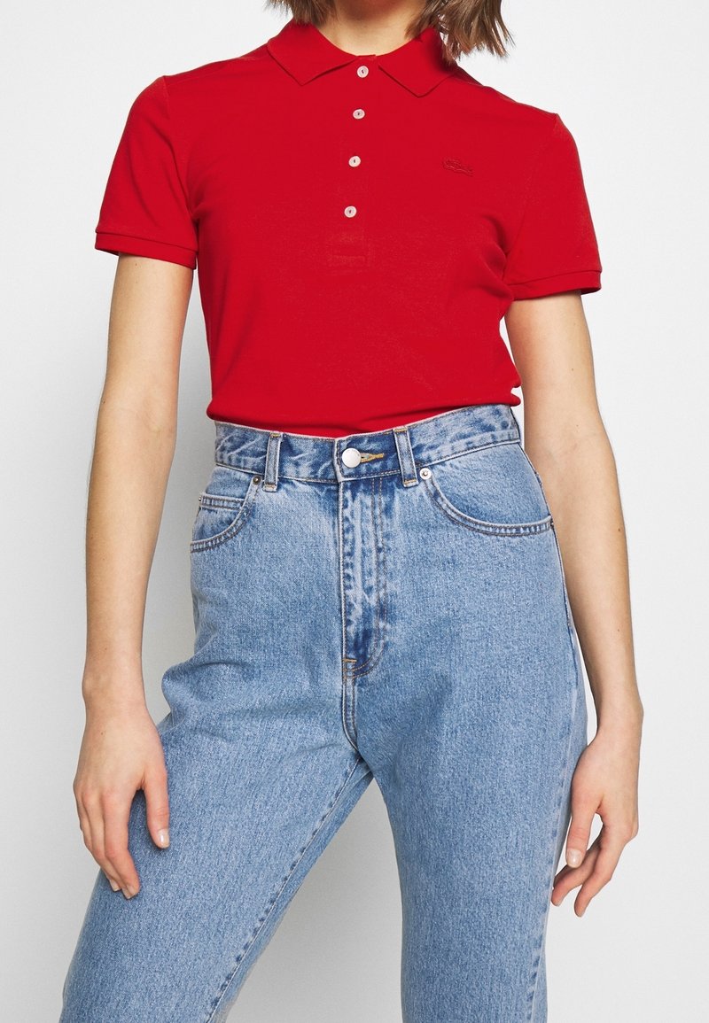 Femme portant un polo rouge ajusté rentré dans un jean en denim bleu clair taille haute avec des poches avant et une fermeture par boutons.