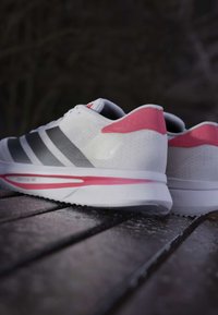 Par hvide og pink løbesko med sorte striber placeret på en træoverflade mod en mørk baggrund.