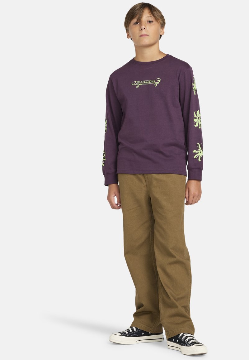 Lila långärmad sweatshirt med grön grafisk design på framsidan och ärmarna, tillsammans med bruna byxor och svarta sneakers.