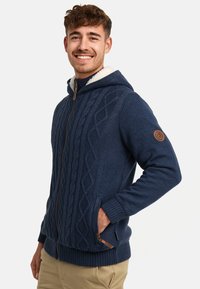 Navy blue knitted hoodie with a cable knit pattern, zip front, and fleece-lined hood. Features a leather logo patch and side pockets.