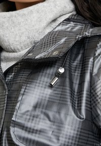 Voděodolná černá kostkovaná bunda na zip s vysokým límcem, stříbrnými patentními knoflíky a elastickým stahovacím šňůrkou pro těsné přizpůsobení. Pod bundou měkký šedý svetr.