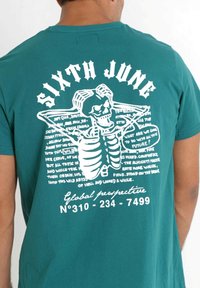 Tyrkysové bavlněné tričko s velkým bílým grafikou kostlivce a textem, včetně "SIXTH JUNE" a "Globální perspektiva" na zadní straně.