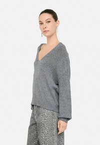 Pull en maille grise avec un col en V, une coupe oversize et des manches longues. La texture est douce, avec de légères taches, associé à un pantalon gris à motifs.
