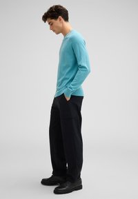 Ein hellblaues, langärmeliges Sweatshirt mit gerippten Bündchen, kombiniert mit lockeren, schwarzen Cargo-Hosen und schwarzen Schuhen, von der Seite betrachtet.