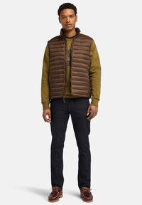 Bruine gewatteerde vest over een olijfgroen sweatshirt, donkere spijkerbroek en bruine boot schoenen met veters, tegen een witte achtergrond.